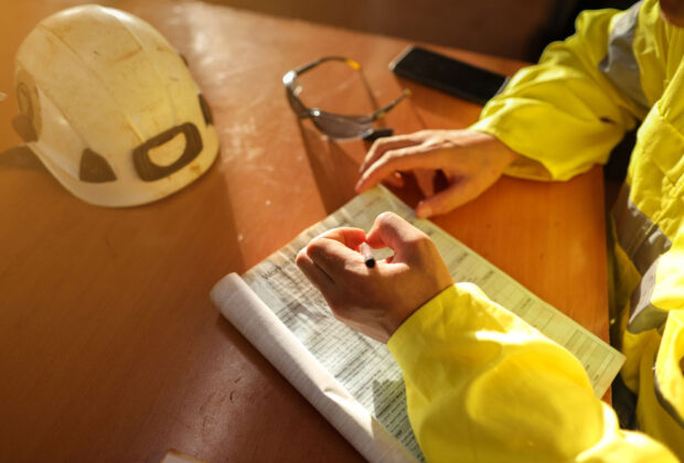 Construction worker in a high-visibility jacket writing on a safety checklist at a desk, with a hard hat and safety glasses nearby.