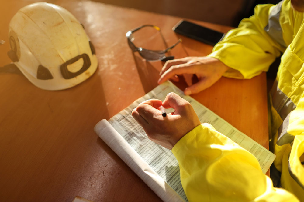 Construction worker in a high-visibility jacket writing on a safety checklist at a desk, with a hard hat and safety glasses nearby.