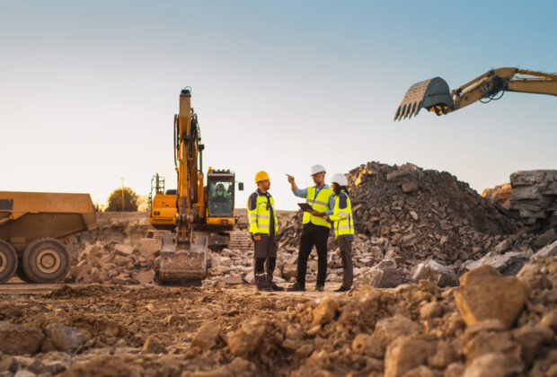 Supervisors and workers in safety gear discussing plans on a construction site with excavators and heavy equipment in the background.