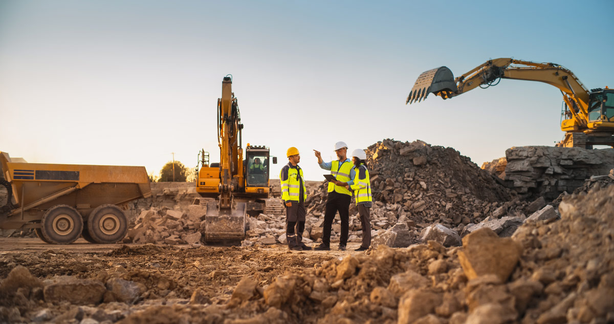 Supervisors and workers in safety gear discussing plans on a construction site with excavators and heavy equipment in the background.